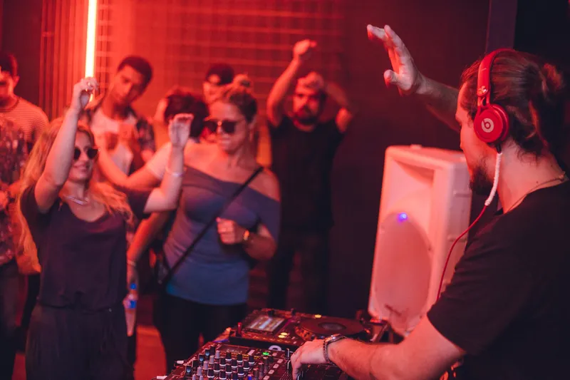 Red-lit dance floor at Vortex — the DJ in red headphones raises a hand while the crowd dances.